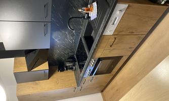 A modern kitchen with wooden cabinets and a black countertop. There is a stove, a sink, and a coffee machine.