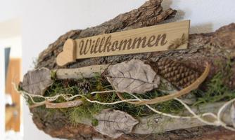 A decorative welcome sign made of wood with natural elements like leaves and pine cones. The rustic design conveys an inviting atmosphere.