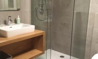 A modern bathroom with a glass shower cubicle and a stylish sink. The walls are in neutral gray tones, and there is a bright wood shelf.