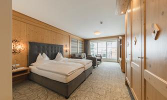 A cozy hotel room with wooden walls and a large window. There is a double bed as well as a seating area with an armchair.