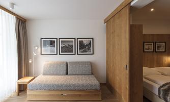 A modern room with a cozy sleeper sofa and wooden accents. Pictures hang on the wall, and there are large windows with curtains.