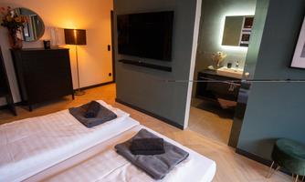 A modern hotel room with two single beds and a television on the wall. The bathroom is visible through a door and features stylish furnishings.