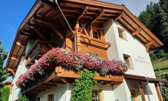 A cozy three-story house with wooden siding and colorful flower boxes. The garden is well-maintained and the sky is blue.