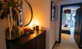 A stylish hallway with a decorative sideboard, a lamp, and a round mirror. A bedroom is visible in the background.