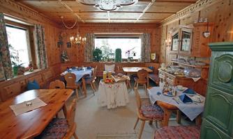 A cozy room with wooden walls and tables. There are chairs, a cabinet wall, and windows with a view outside.