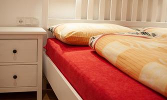 A cozy bedroom with a red bed sheet and two pillows. Next to the bed stands a small nightstand.