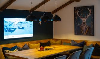 A cozy room with a large wooden table and comfortable cushions. A picture of a stag hangs on the wall, and a screen displays a snowy landscape.