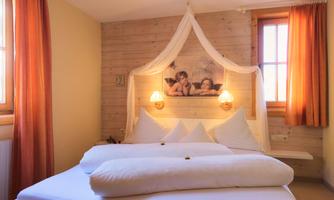 A cozy bedroom with a large bed and decoration in warm colors. The walls are made of wood, and large windows let in plenty of light.