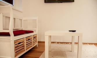 A bright room with a white table and a bench. On the bench lies a red cushion and there is a television on the wall.