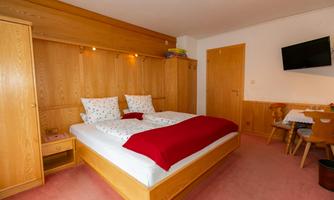 A cozy bedroom with a double bed and wooden furniture. A television hangs on the wall, and there is a small table with chairs.