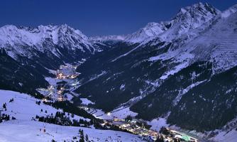 A snowy mountain landscape at night with sparkling lights in the valley. The snow-covered peaks and the clear sky give the scene a tranquil atmosphere.