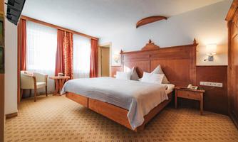 A cozy hotel room with a large bed, a wooden bedside table, and a small table. The walls are painted in warm colors, and large windows bring in daylight.
