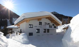 A modern chalet in the snow, surrounded by mountains. The sun shines clearly over the building.