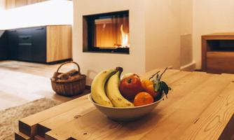 A bowl of fresh fruits like bananas, apples, and oranges is on a wooden table. In the background, a fireplace is burning, creating a cozy atmosphere.