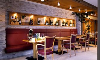 A stylish restaurant with red upholstered benches and wooden chairs. In the background, wine bottles are decorated on a shelf.