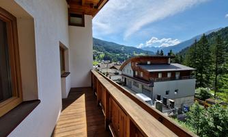 A balcony with a wooden railing that offers a view of the mountains and surrounding houses. The sky is clear and the landscape is green.