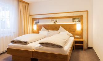 A cozy bedroom with a wooden double bed and two pillows. The room is bright and friendly with a large window and a simple, modern design.