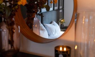 A stylishly decorated room with a round mirror that shows a cozy bed and flowers. A burning candle creates a warm atmosphere.