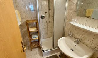 A modern bathroom with a shower and a sink. The walls are tiled in light colors and there is a shelf with towels.