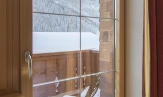 A cozy balcony with a wooden chair overlooking a snowy landscape. The view includes mountains and a clear winter sky.
