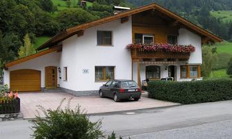 A cozy house in alpine style with a large garden and a driveway. Colorful flowers adorn the windows and the surroundings are surrounded by green hills.