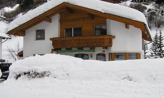 A cozy house with wooden paneling, surrounded by a thick layer of snow. In the background, snow-covered mountains can be seen.