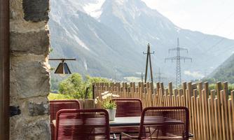 A terrace with red chairs and a table, surrounded by a breathtaking mountain landscape. In the background, tall mountains and a clear sky can be seen.