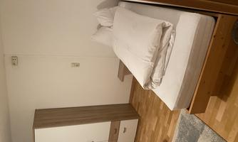 A simple bedroom with a single bed and a dresser. The floor is wooden and the walls are painted light.