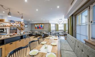A modern restaurant with wooden furniture and a cozy atmosphere. Tables are set for guests, and there is a large window with plenty of natural light.