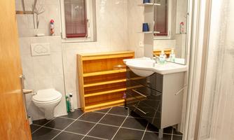 A modern bathroom with a shower, a sink, and a wooden shelf. The walls are light tiled and the floor is dark tiled.