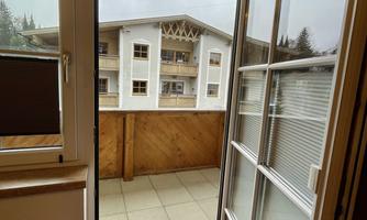 A balcony with a view of a building. The windows are open and there is a carpet in the room.