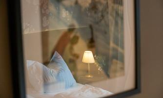 A cozy bedroom with a soft bed and a nightstand lamp. In the background, a decorative wall design is visible.