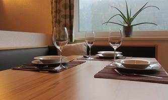 A table decorated with dishes and glasses, ready for a meal. In the background, there is a plant and a large window view.