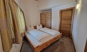 A cozy bedroom with a wooden bed and two pillows. The walls are light, and there is a window with curtains.