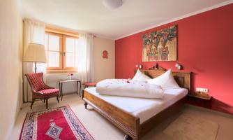 A cozy bedroom with a large bed and red wall color. There is a table, a chair, and decorative art on the wall.