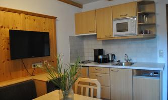 A modern kitchen with wooden shelves and appliances such as a microwave and kettle. A dining table with chairs and a television on the wall complete the cozy atmosphere.