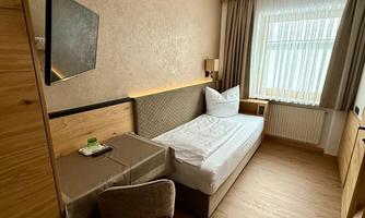 A simple hotel room with a single bed and wood paneling. There is a desk and a window with curtains that lets in natural light.