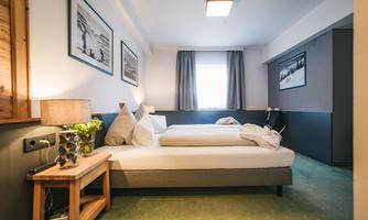 A modern bedroom with two single beds and light curtains. Stylish pictures hang on the wall, and a bedside table with a lamp and a plant stands next to the beds.
