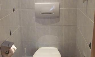 A small toilet with white tiles and a modern toilet. The amenities include toilet paper and a trash bin.