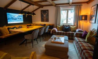 A cozy living room with a wooden beam ceiling and modern furniture. There is a large table, a comfortable couch, and large windows that let in light.
