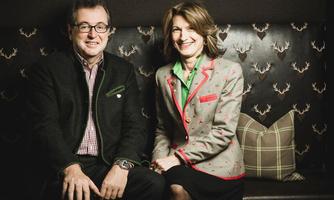 A pair of people is sitting on a couch with a stylish, patterned background. Both are smiling and wearing fashionable, formal clothing.