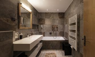 A modern bathroom with a bathtub, a sink, and a large mirror. The walls are decorated with gray tiles and there is a towel heater.