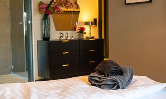 A modern bedroom with a cozy bed and freshly folded towels. In the background, there is an elegant dresser with decorations.