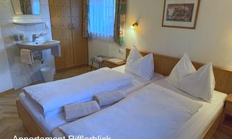A cozy bedroom with two single beds and white pillows. In the background, there is a sink and a window through which daylight enters.