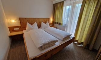 A cozy bedroom with a double bed and two pillows. Large windows with curtains let in plenty of daylight.