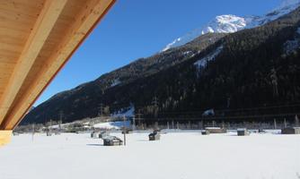 A snowy valley with mountains in the background and a clear blue sky. In the foreground, there are empty fields and a few small structures visible.