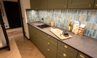 A modern kitchen with green cabinets and an elegant countertop. In the background, blue tiles and a kitchen unit with accessories are visible.