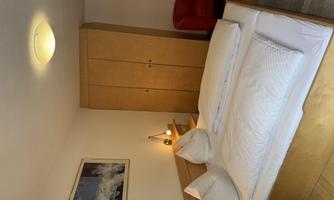A simple bedroom with a double bed, white pillows, and a bright ceiling lamp. A red armchair and a wooden wardrobe complete the room.