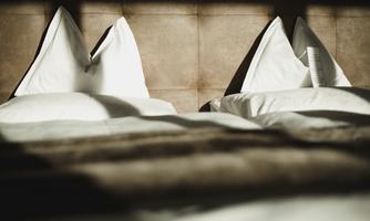 A cozy bed with white pillows and a soft blanket. Gentle light falls on the bedding, creating a relaxed atmosphere.