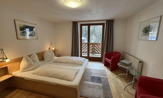 A cozy bedroom with a double bed and two red armchairs. Large windows offer a view of the balcony.